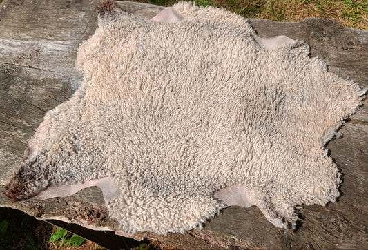 A natural sheepskin or hide rug with curly off-white and light tan fur, laid flat on a weathered wooden log outdoors.