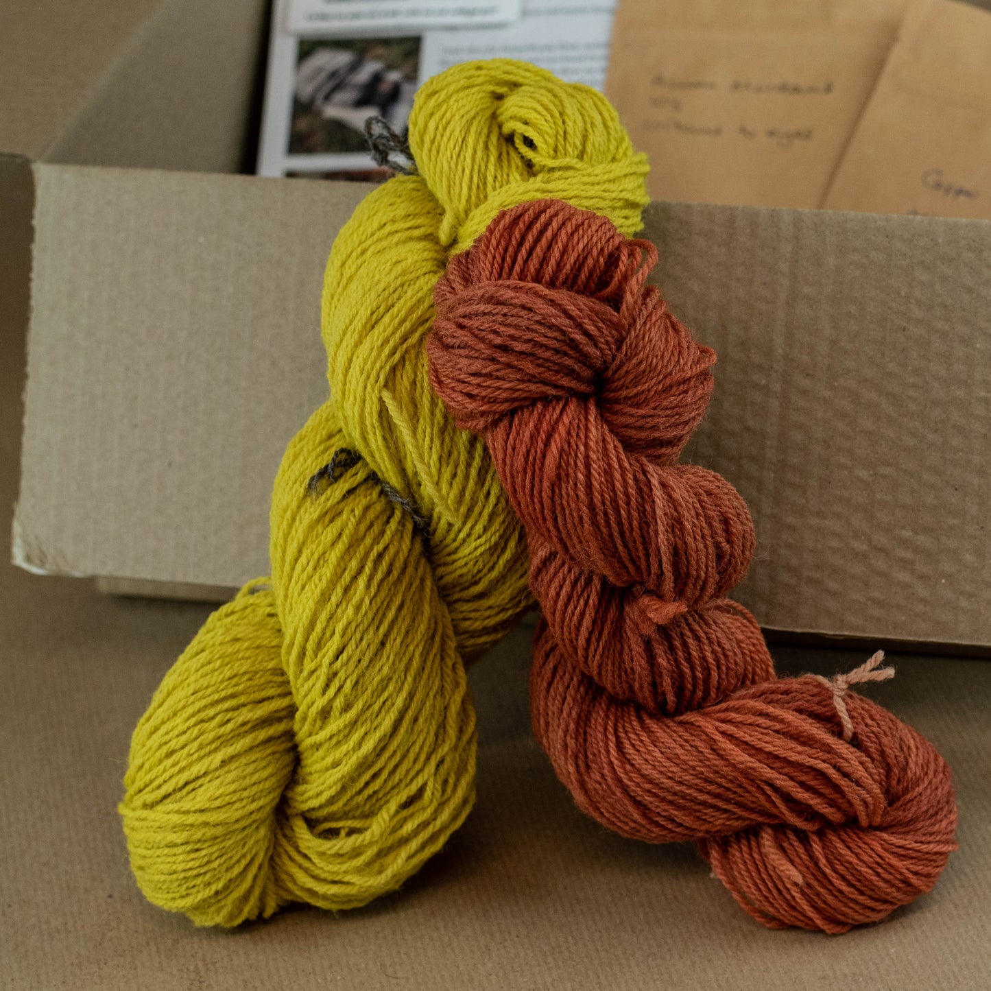 Two skeins of yarn, one yellow and one red-brown, leaning against a cardboard box on a brown surface.