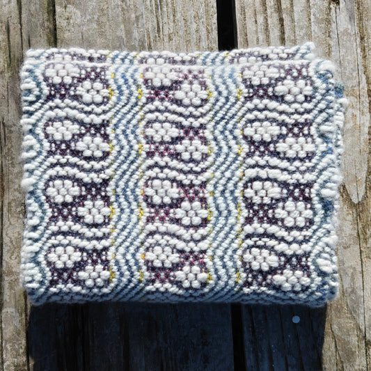 A folded scarf with white hand spun lambswool in a pattern like paw prints with lines circling around them, on a yello, blue and purple background.
