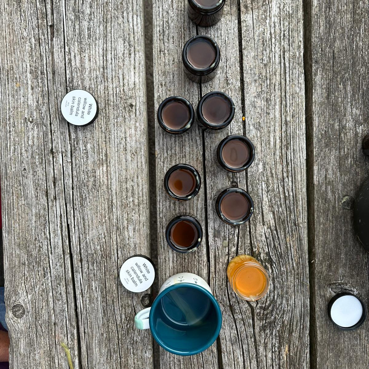 Assorted small containers of freshly made willow balm, cooling, and a blue mug on a wooden surface
