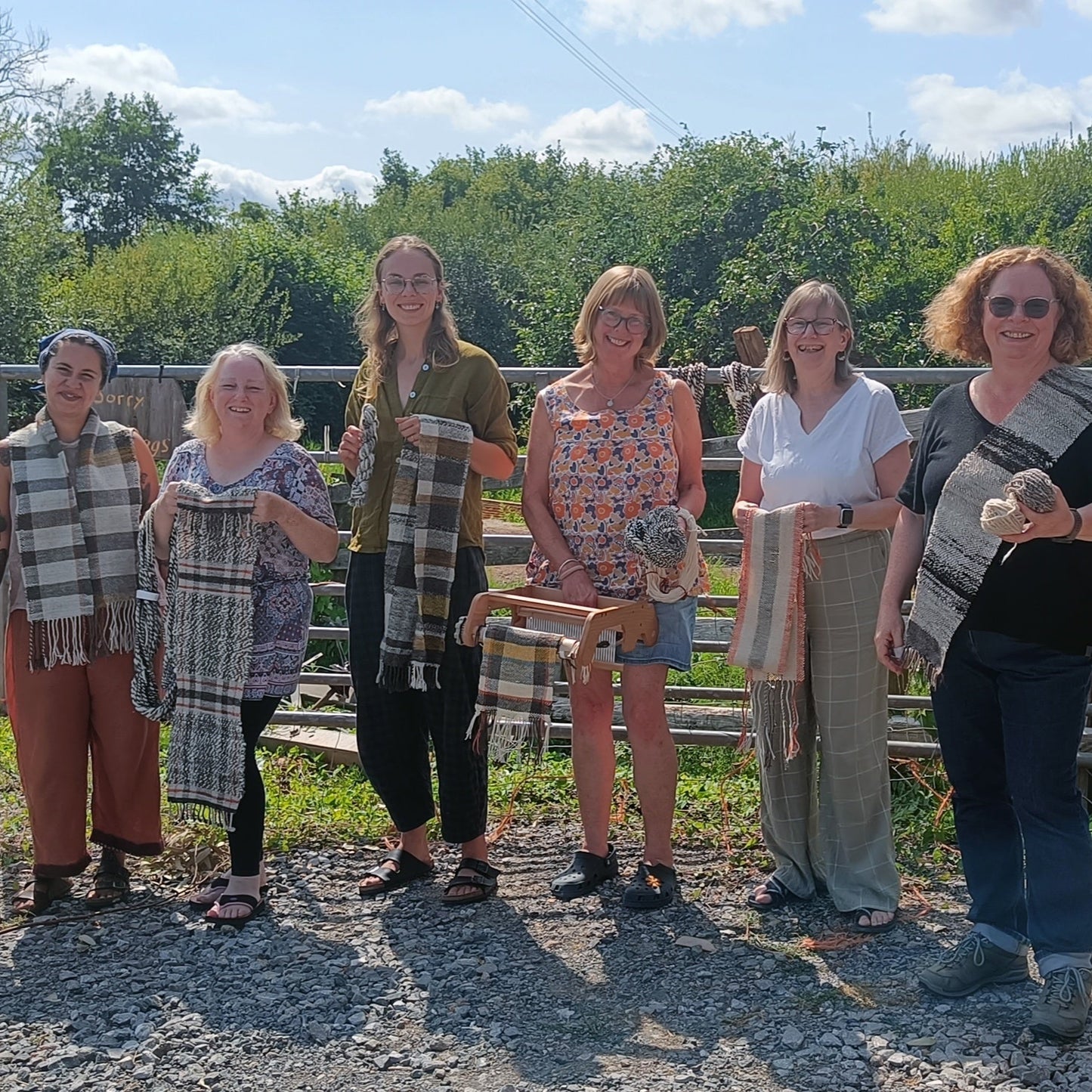 Group of people standing outdoors in a rural setting with greenery and a wooden fence.