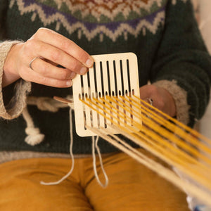 Image shows a pair of hands using the band weaving set, with the heddle raised and orange threads lifted.
